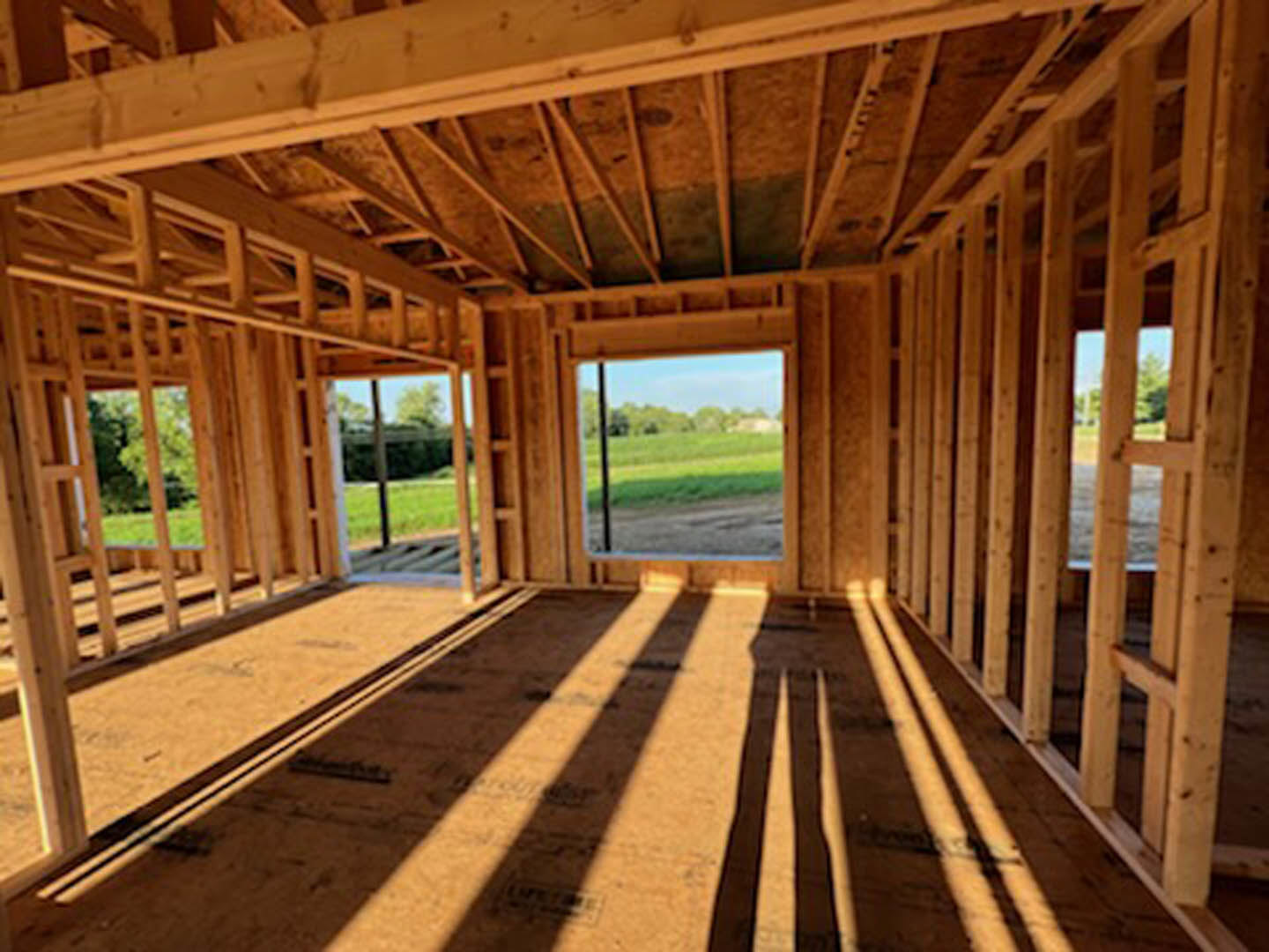 Sunlit room with wooden plank flooring, large window overlooking green grass and trees, shadow cast across floor, exposed wood ceiling beams