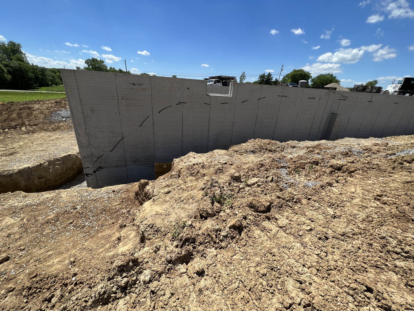 Concrete foundation wall with central opening, large dirt mound in foreground, scattered rocks, white pickup truck parked near unfinished building, surrounding trees, blue sky with