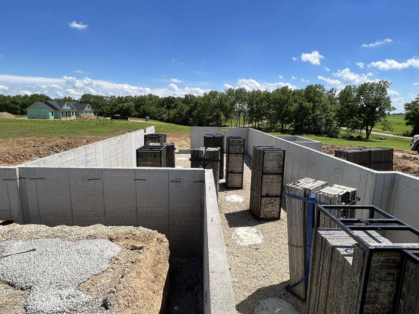 Concrete foundation with stacked wooden boxes, black-framed brick wall, black roof, gravel and rocks piled nearby, sign with black frame, blue sky with scattered clouds and
