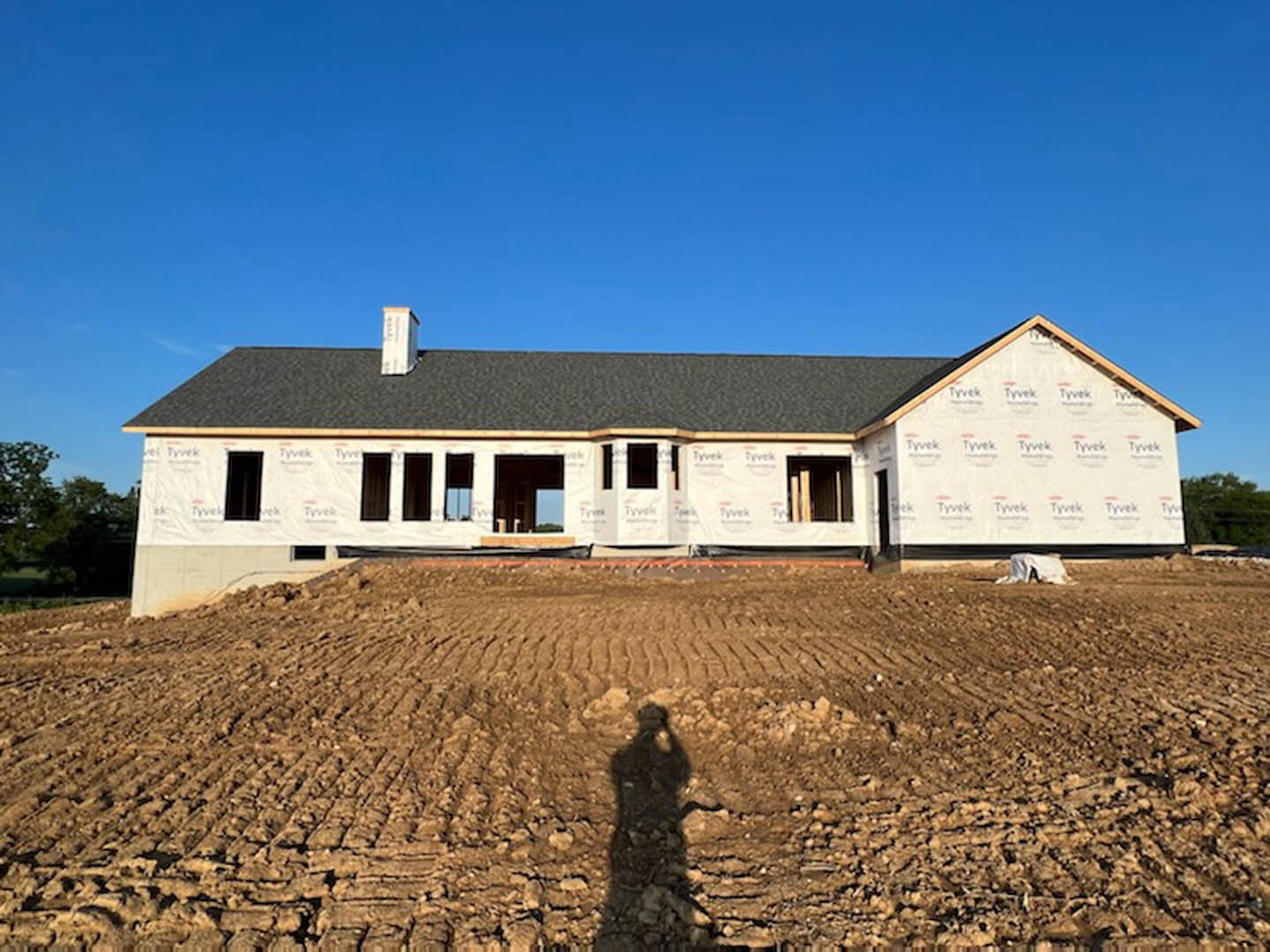 Framed house under construction on dirt lot with exposed wooden beams, blue sky overhead, and tree in background