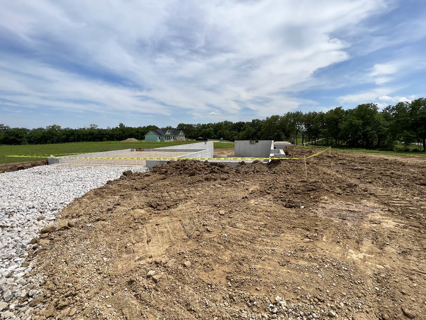 Partially built house with blue roof, exposed framing, dirt and grass lot, yellow caution tape, person standing near unfinished wall, blue sky with scattered clouds