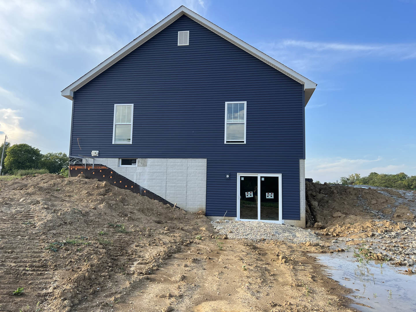 Blue siding home with white-framed windows, glass door displaying a sign, set against a backdrop of a dirt hill and cloudy sky.