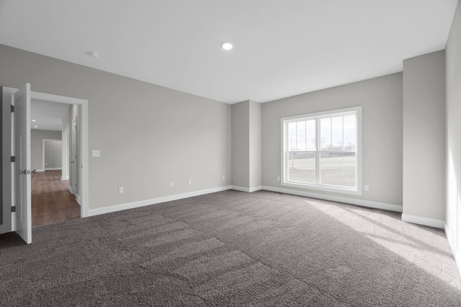Carpeted room with white walls, large window overlooking grassy field, white door, ceiling light fixture, and hallway with matching doors