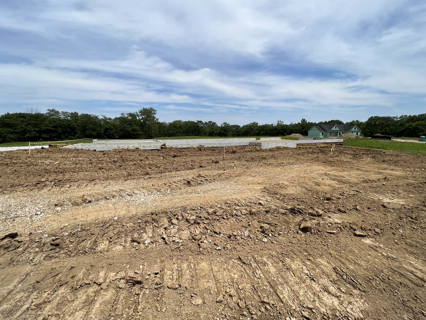Dirt field bordered by a wooden fence and road, blue-roofed house in the distance, group of trees under a partly cloudy sky