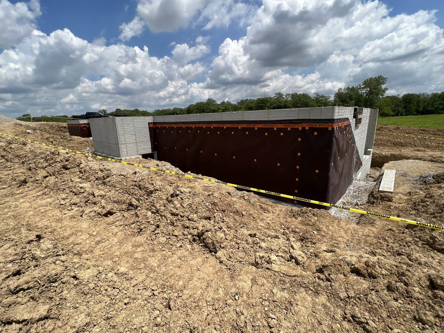 White block wall under construction with barrier tape, dirt pile, and rocky ground; white walkway leads past trees and grass beneath a blue sky with scattered clouds.