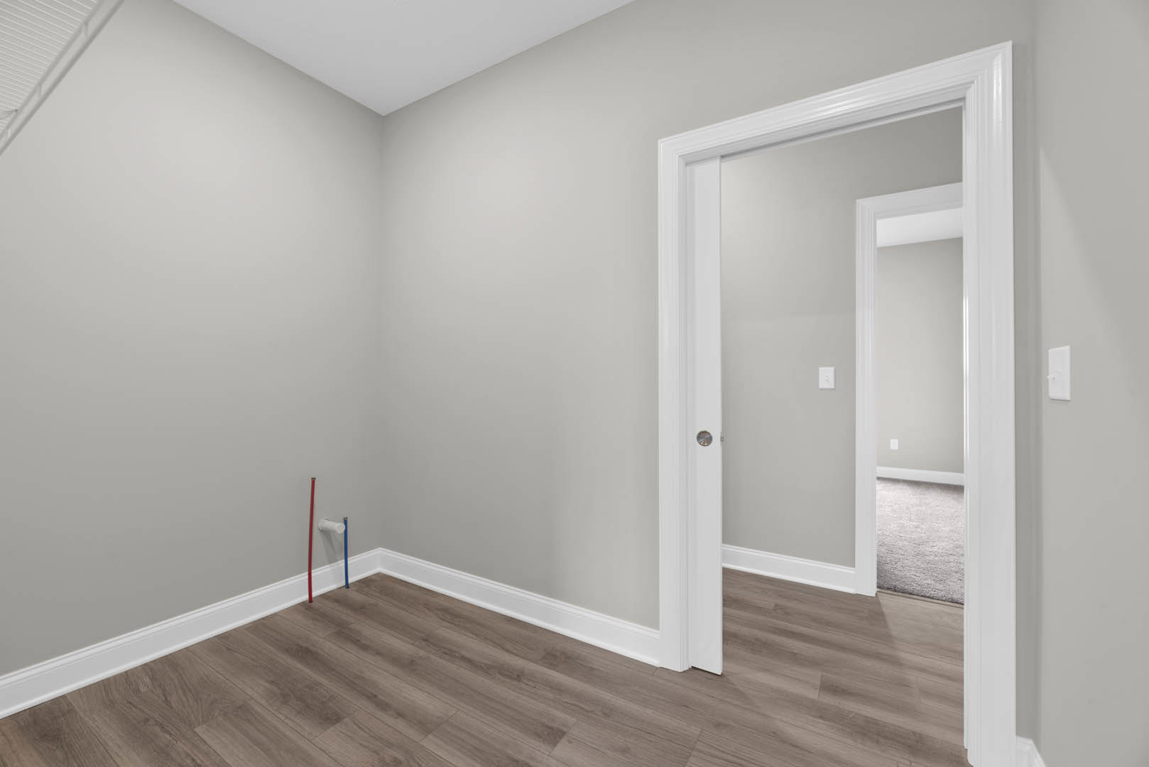 White door open to a room with wood flooring, white baseboards, and grey carpet; white wall and visible red and blue plastic tubes along the floor.