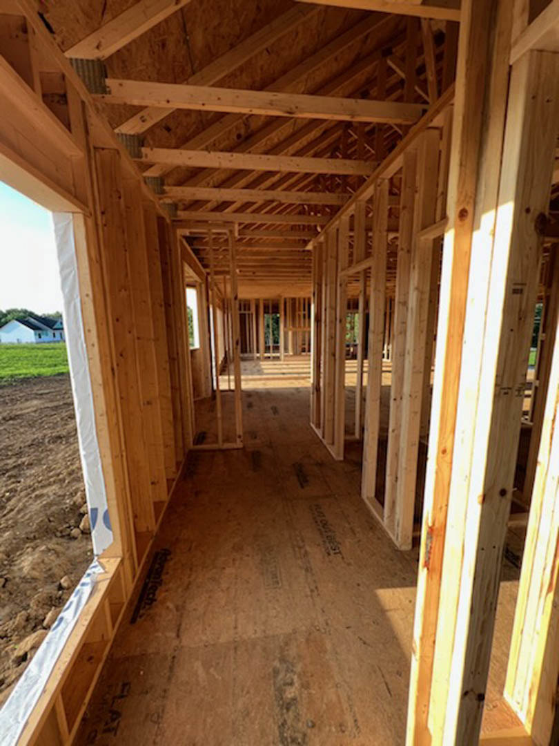 Wood framing and exposed beams forming the structure of a house under construction, with unfinished floors and open window spaces, surrounded by trees in the background.