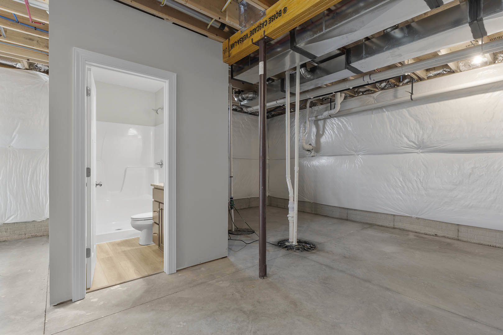 Bathroom with white toilet, walk-in shower, concrete floor, exposed pipes and wires, plaster walls, wooden ceiling beam with black text, and building insulation visible.