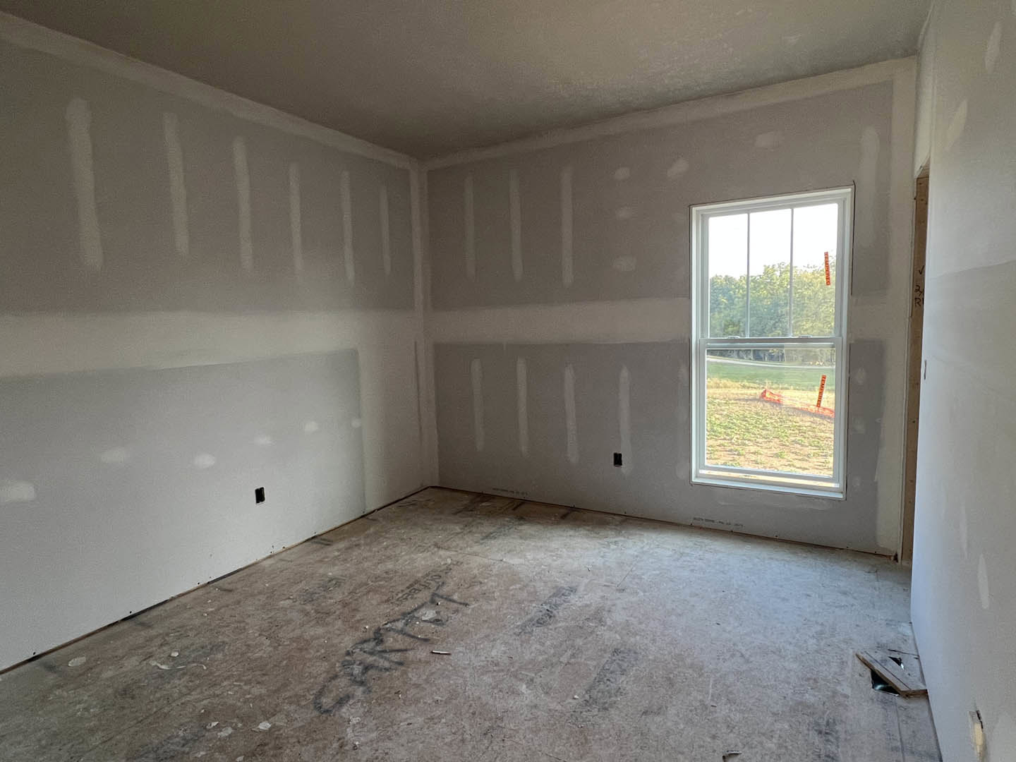 Unfinished room with two white-framed windows, plaster walls showing holes, dirty floor marked with black writing, and a view of a fenced field outside