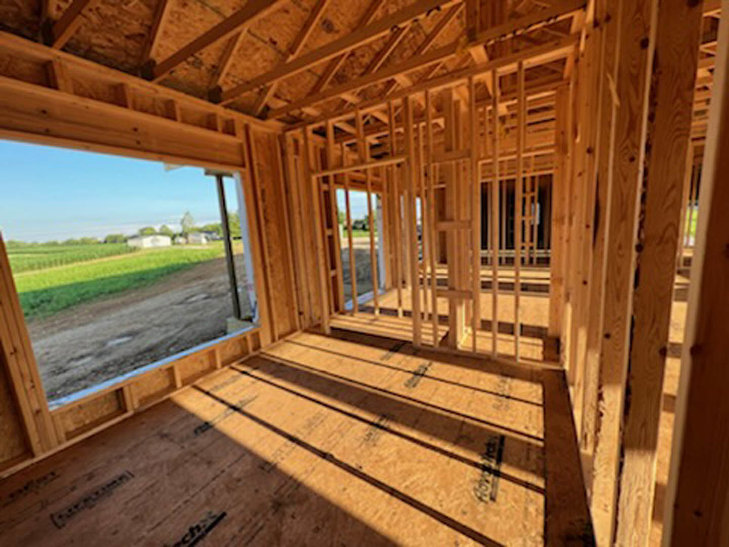 Framed interior with exposed wooden beams, unfinished plywood floor marked with black lines, large window letting in natural light