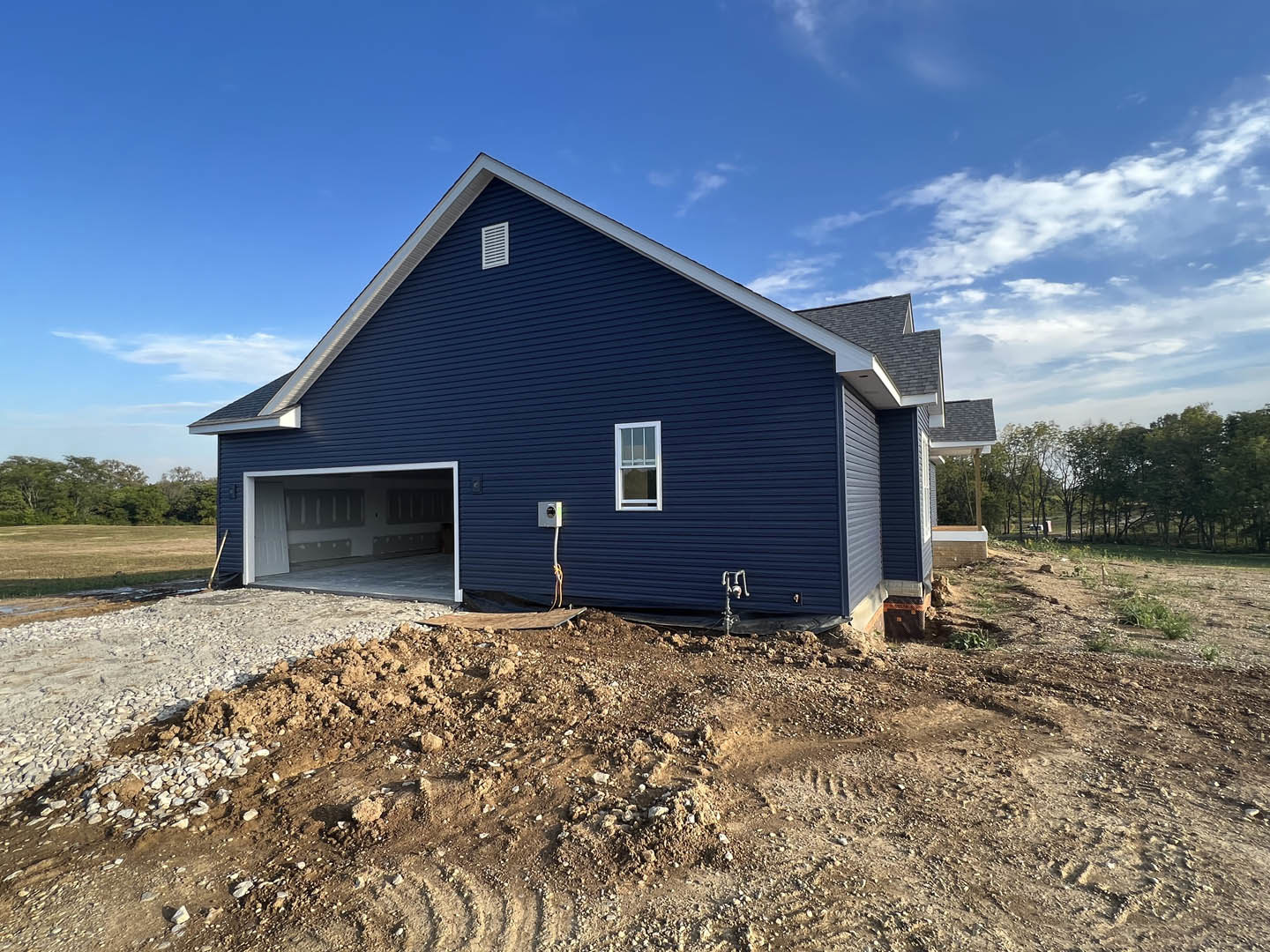 Blue house with white roof and white-framed windows, attached garage with closed door, surrounded by dirt lot and mature trees under partly cloudy sky
