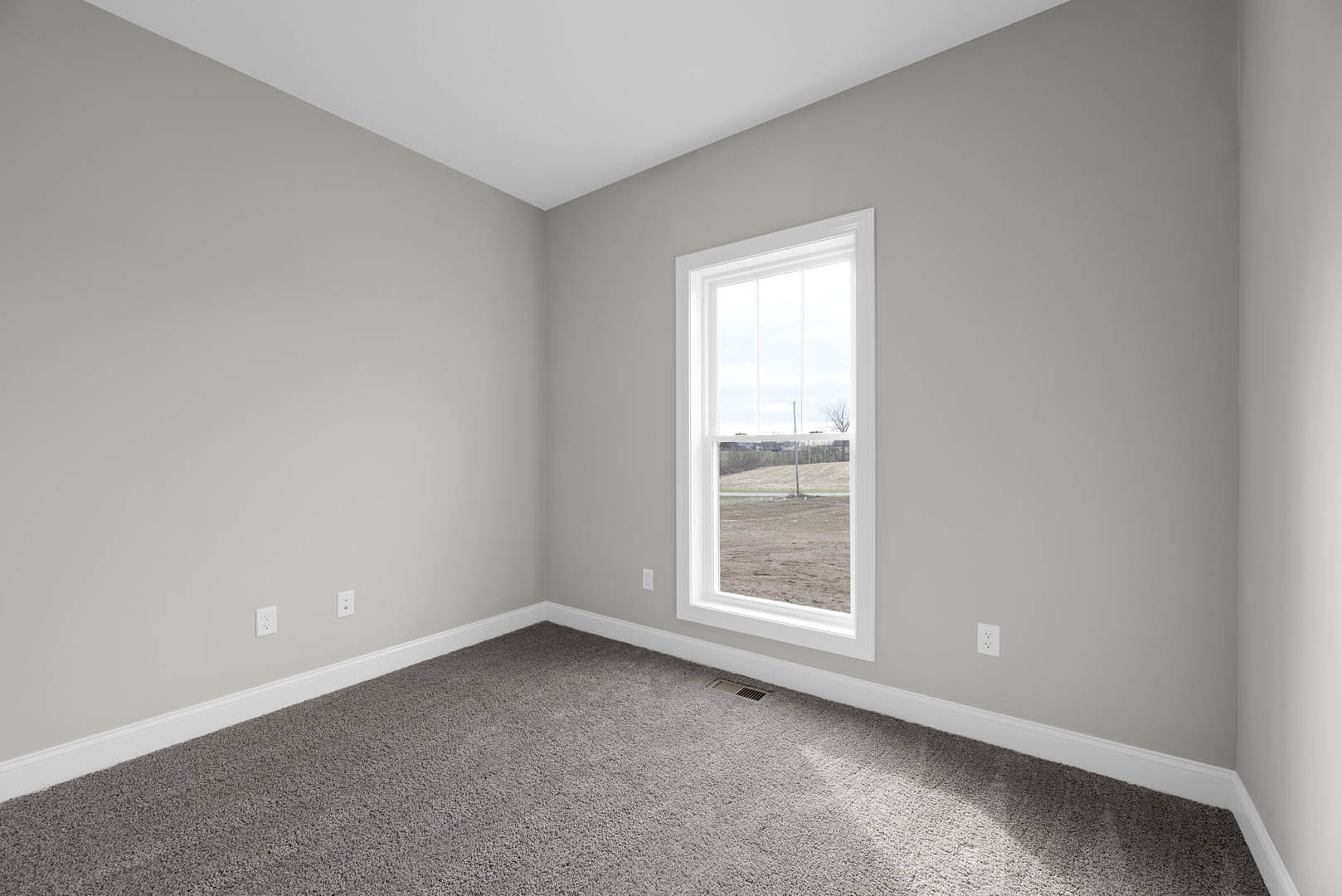 Bright room with white walls, large window framed in white overlooking a dirt field, grey carpeted floor with vent in corner, natural light casting shadows