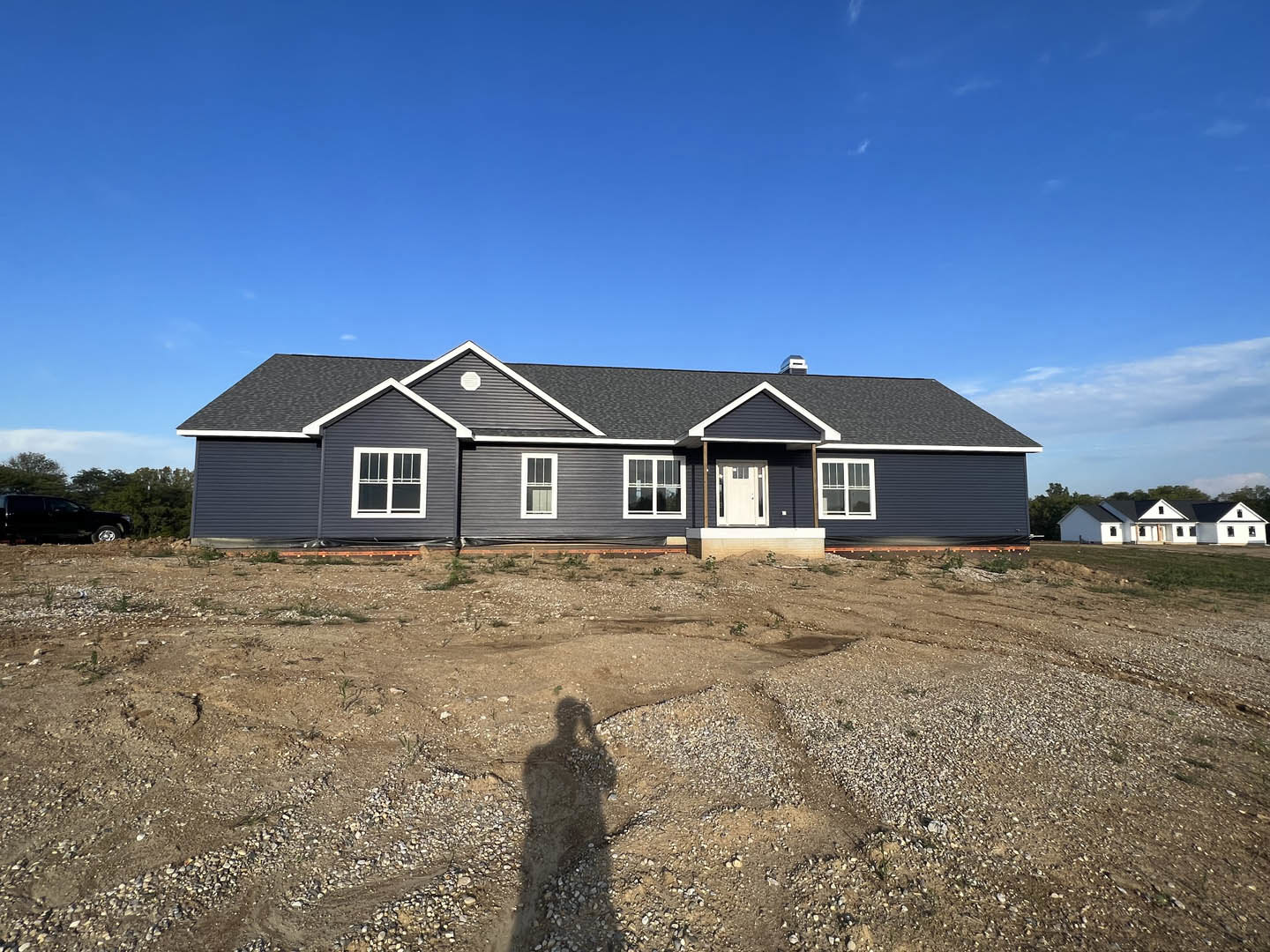 White house with black roof and covered porch, set on a dirt field under a blue sky with scattered clouds
