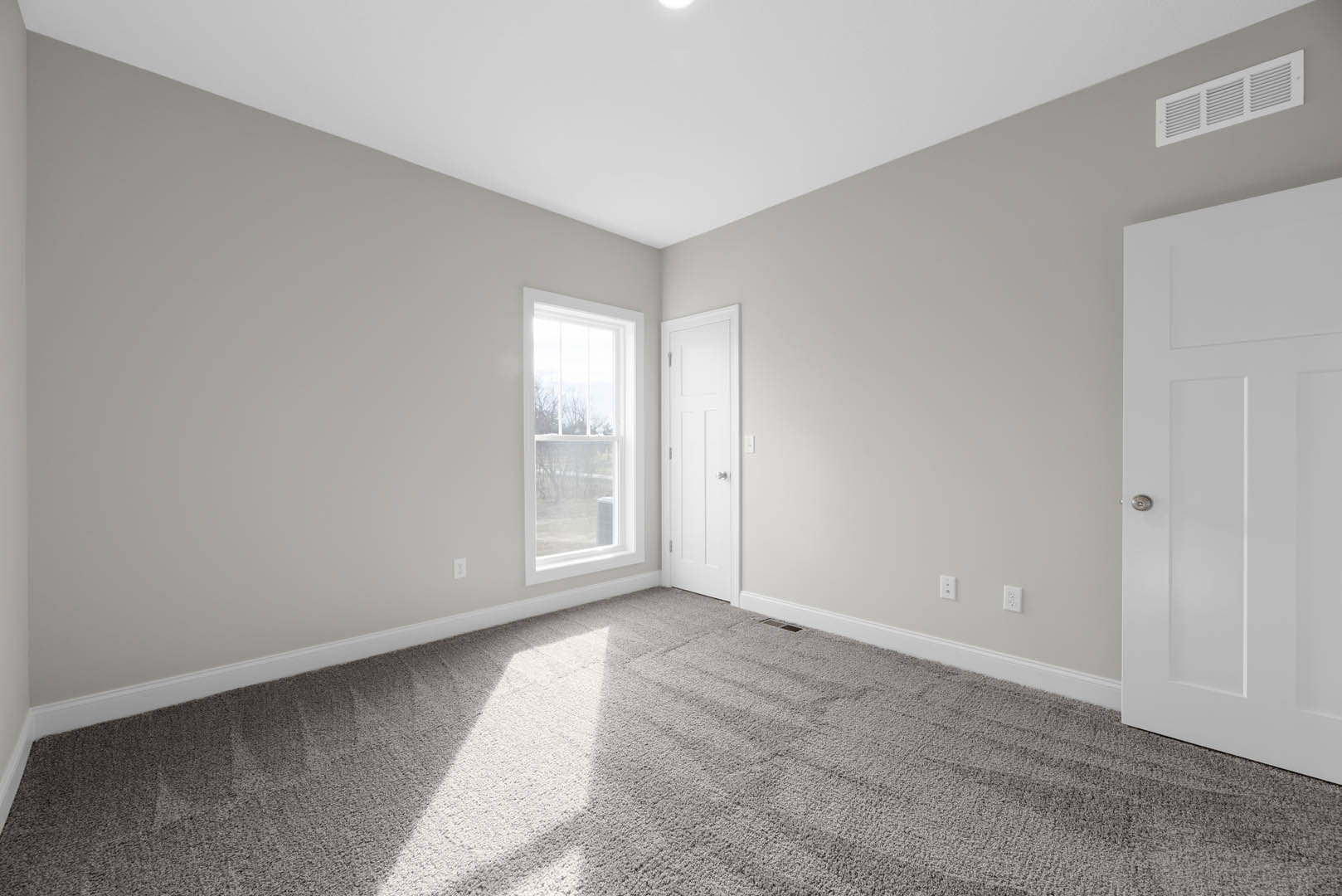 White paneled door with silver knobs, beige carpet flooring, large window overlooking trees, white walls, ceiling vent