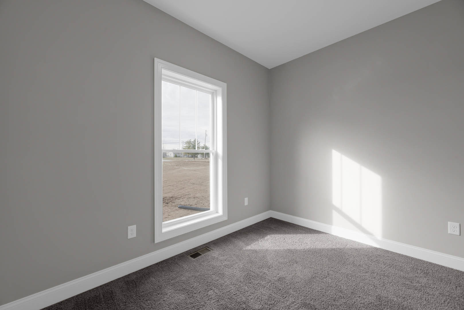 Grey carpeted floor with a rectangular vent in the corner, white electrical outlet on a plain wall, large window overlooking a dirt field, simple baseboard molding along the