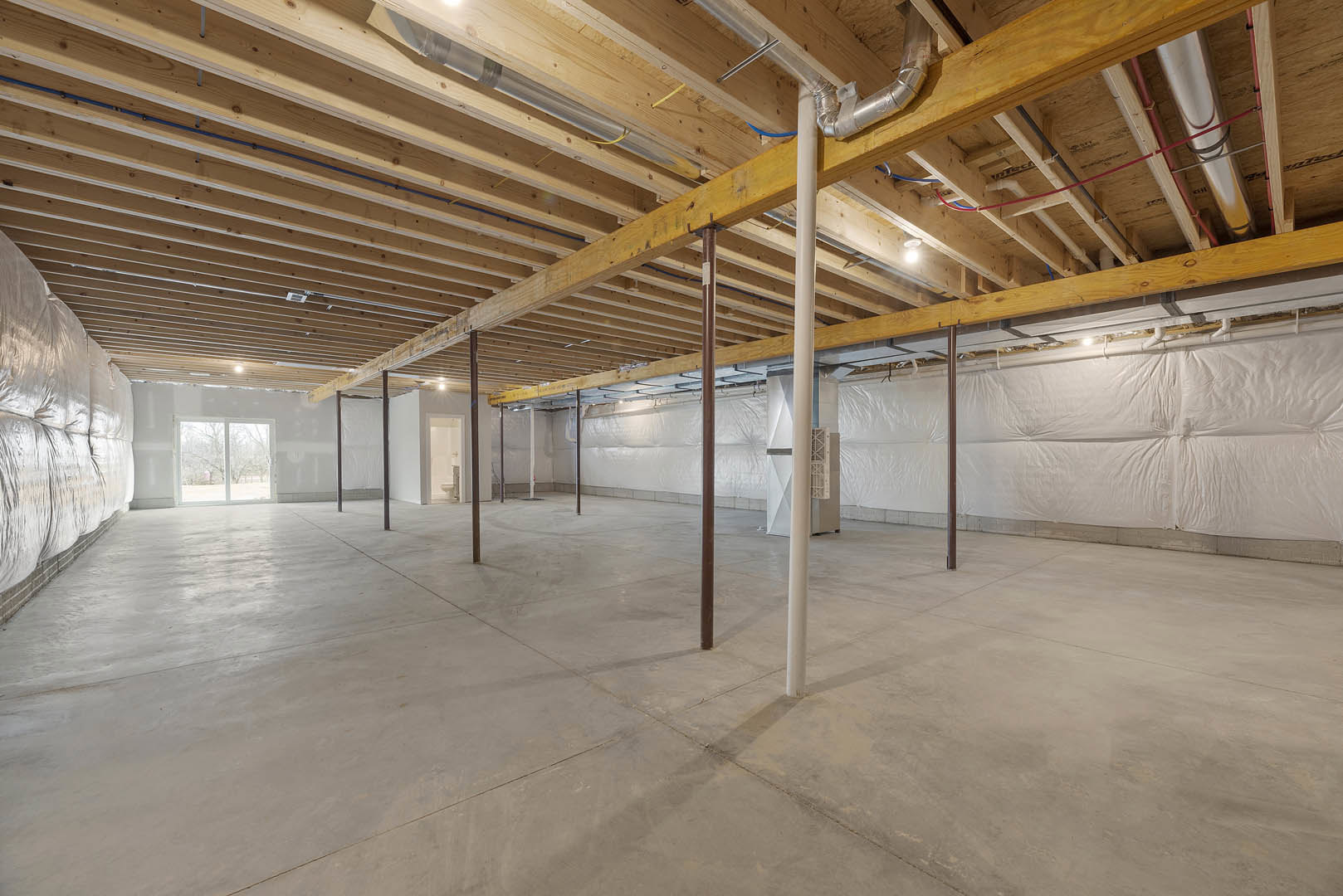 Spacious room featuring exposed wooden beams and ceiling, concrete floor with structural pillars, large window overlooking trees, visible pipes, and a close-up of a white blanket