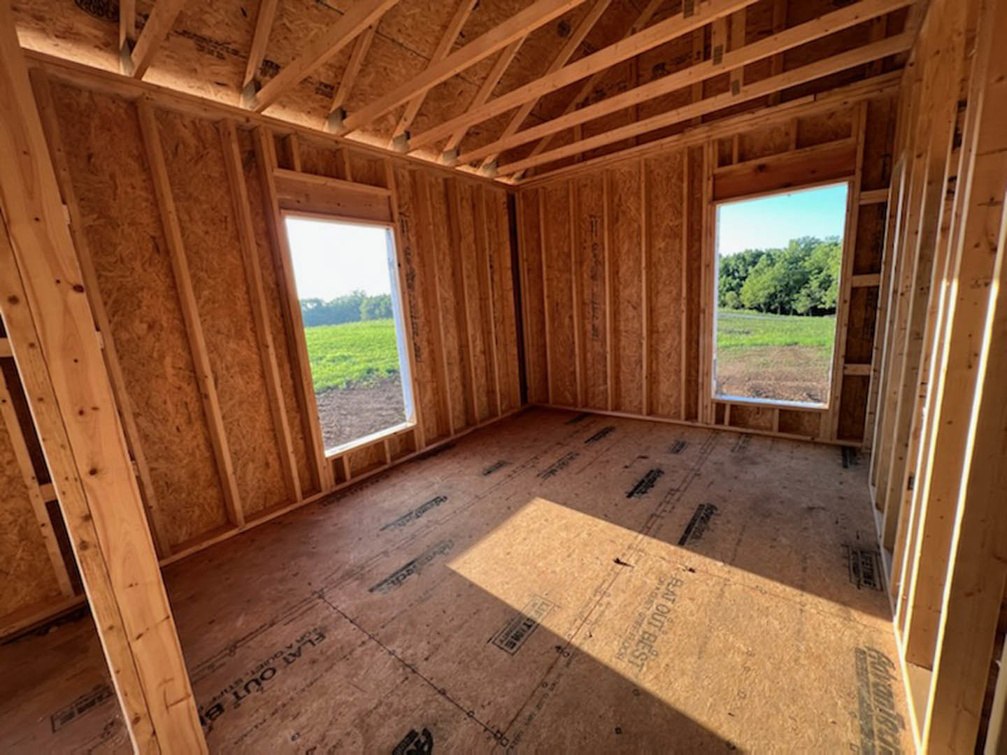 Bright room with large windows overlooking a grassy field, hardwood floors, exposed beams, and plywood accents.