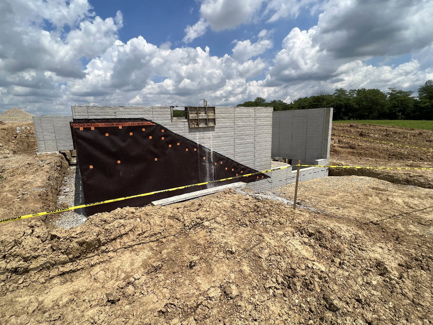 Construction site with wire fencing, brick and concrete walls, black mesh covering, pile of dirt, grassy ground, and blue sky with scattered clouds