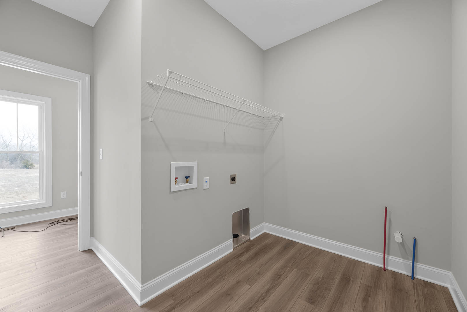 Wood floor room with white plaster walls, white shelf mounted above a wall, white square panel with two valves, window overlooking snowy field, trash can in corner