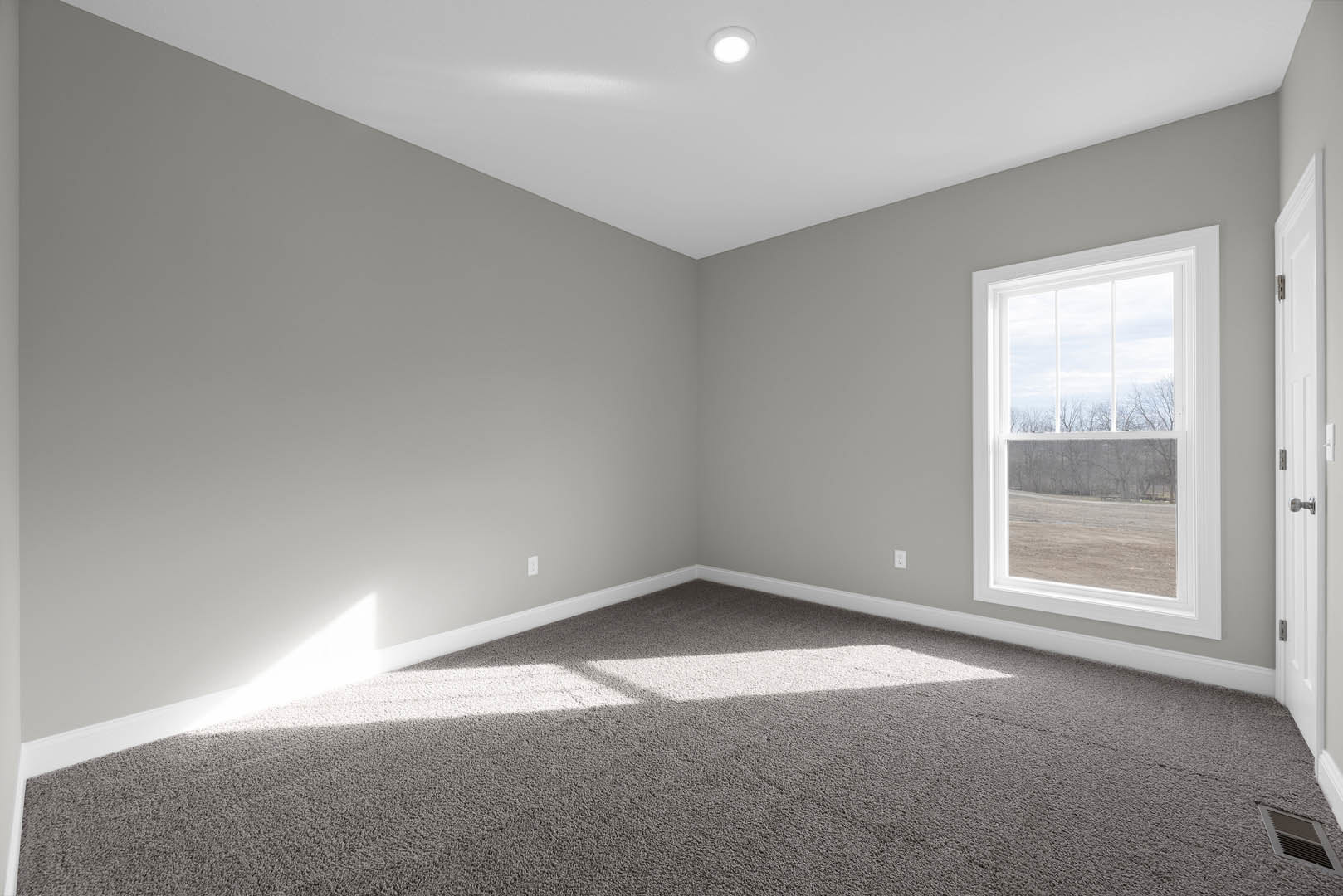Carpeted bedroom with white walls, large window overlooking grassy field, recessed ceiling light, wall vent, and crown molding.