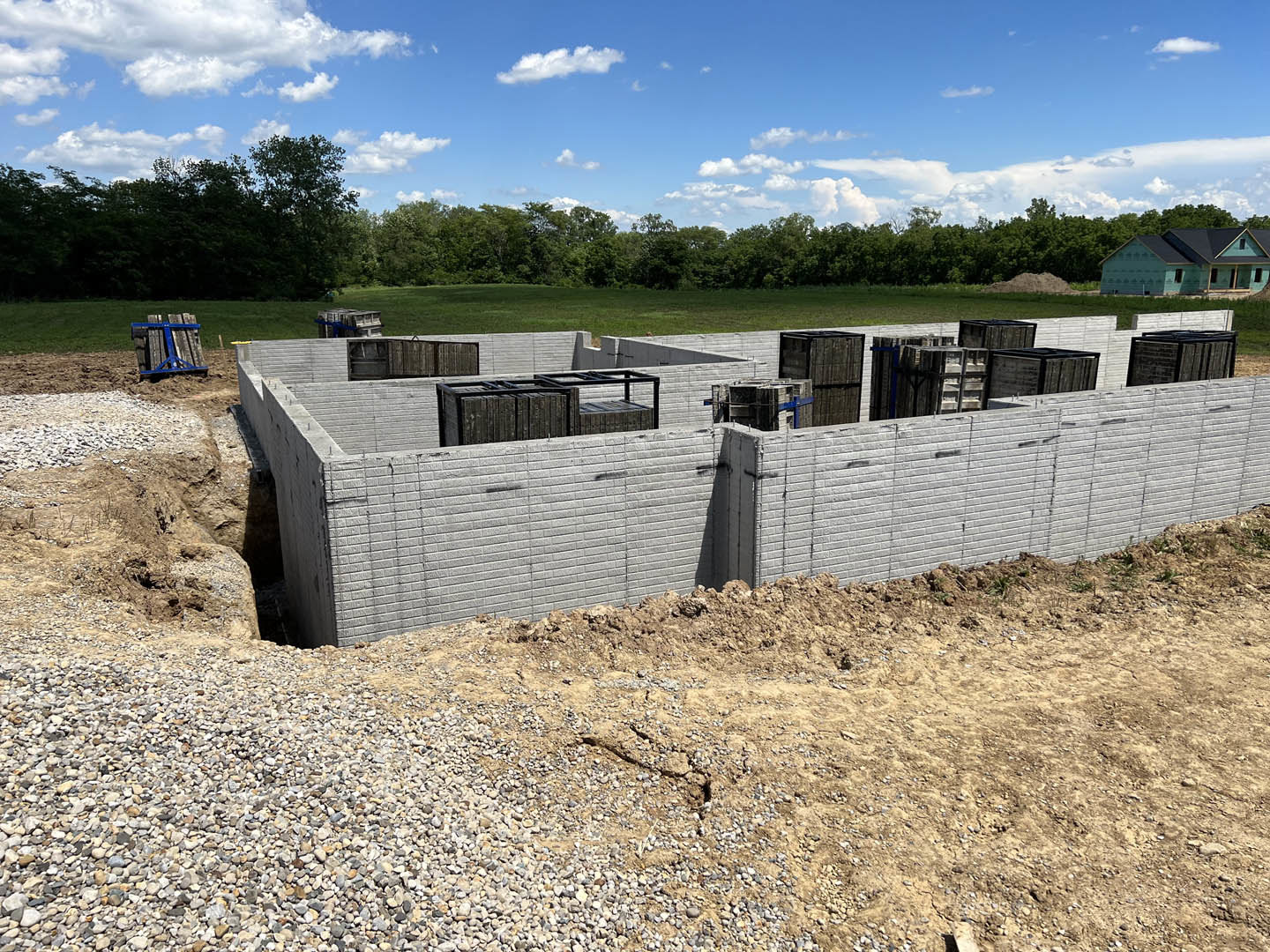 Concrete walls and brick foundation surrounded by piles of wood and dirt, black roof partially installed, blue metal framing visible, construction site bordered by trees under a