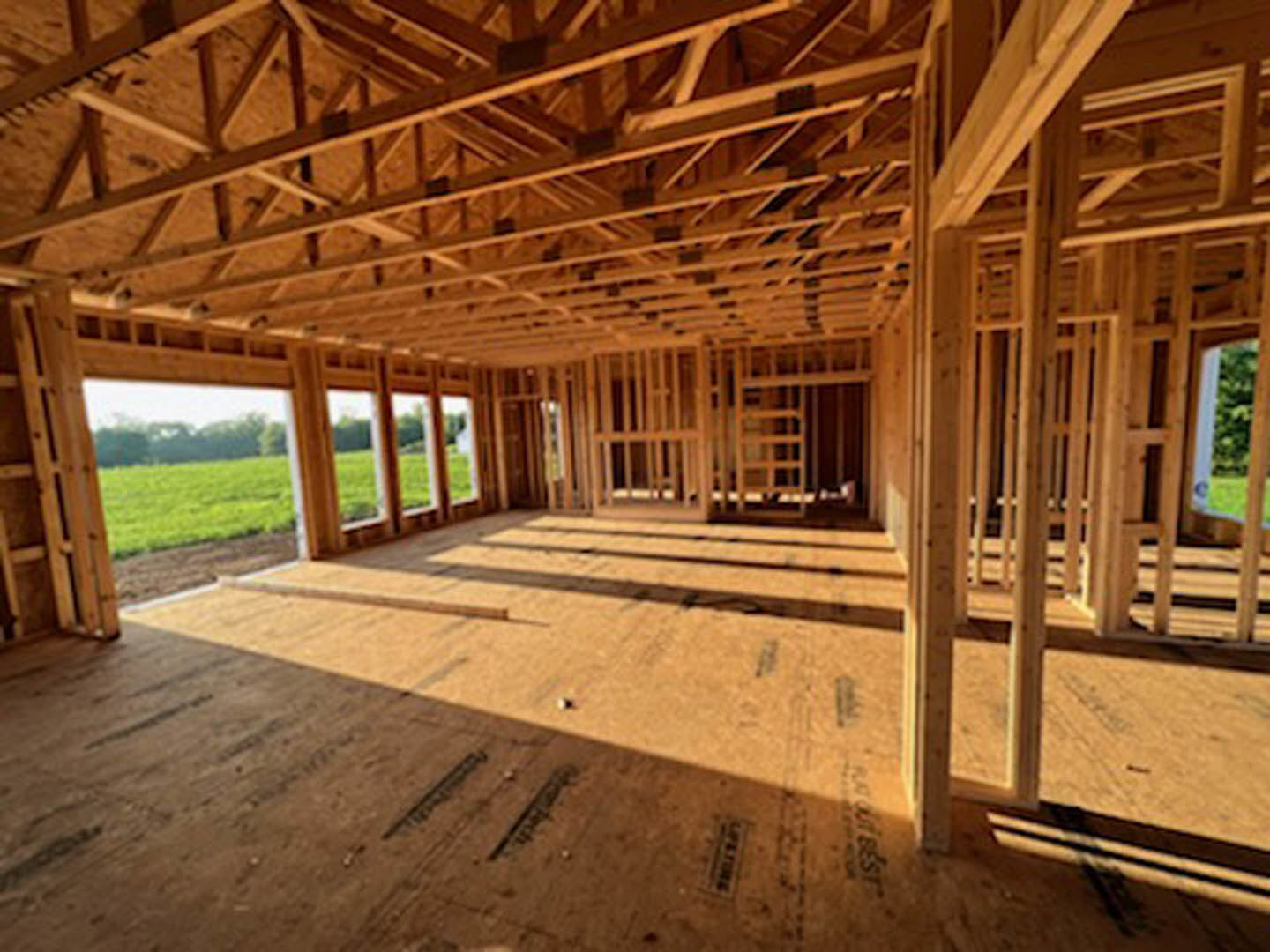Room with exposed wood framing, unfinished plank floor, large windows overlooking grassy field and trees