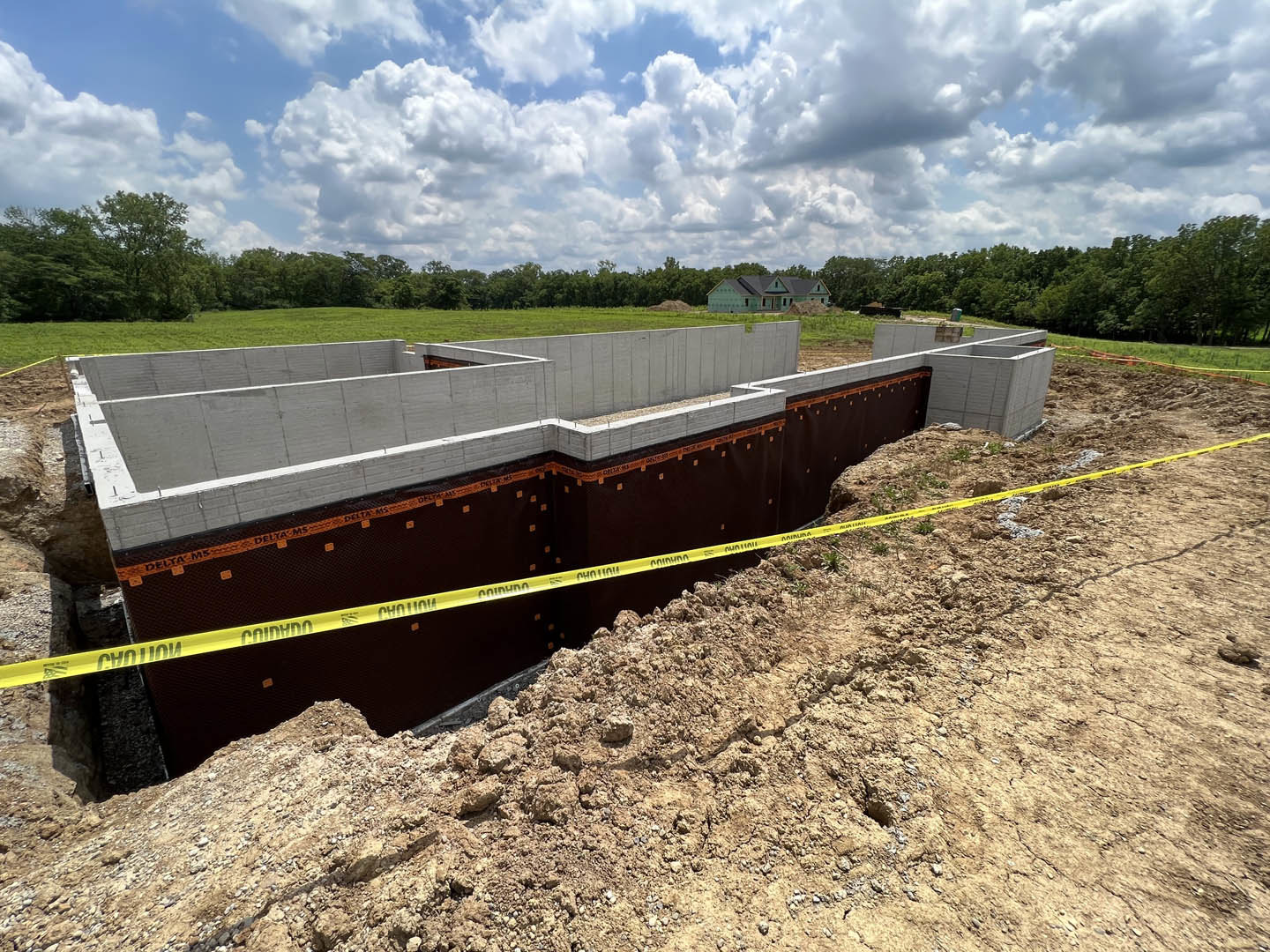 Excavated construction site with exposed concrete foundation, orange and yellow safety tape, grassy ground, partially built house with green roof, blue sky with scattered clouds