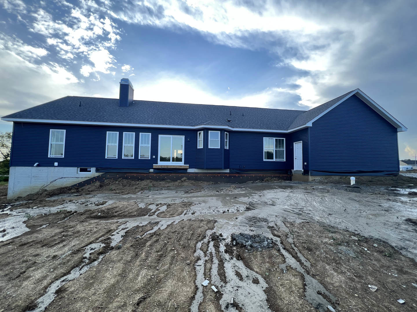 Blue house with white-trimmed windows, blue siding, dirt hill in foreground, muddy area with blue tarp, reflected trees in window, partly cloudy sky overhead