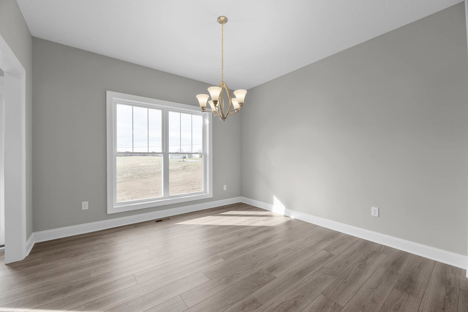Wood flooring in a spacious room with white plaster walls, decorative molding, large window overlooking a grassy field, and a five-light chandelier hanging from the ceiling