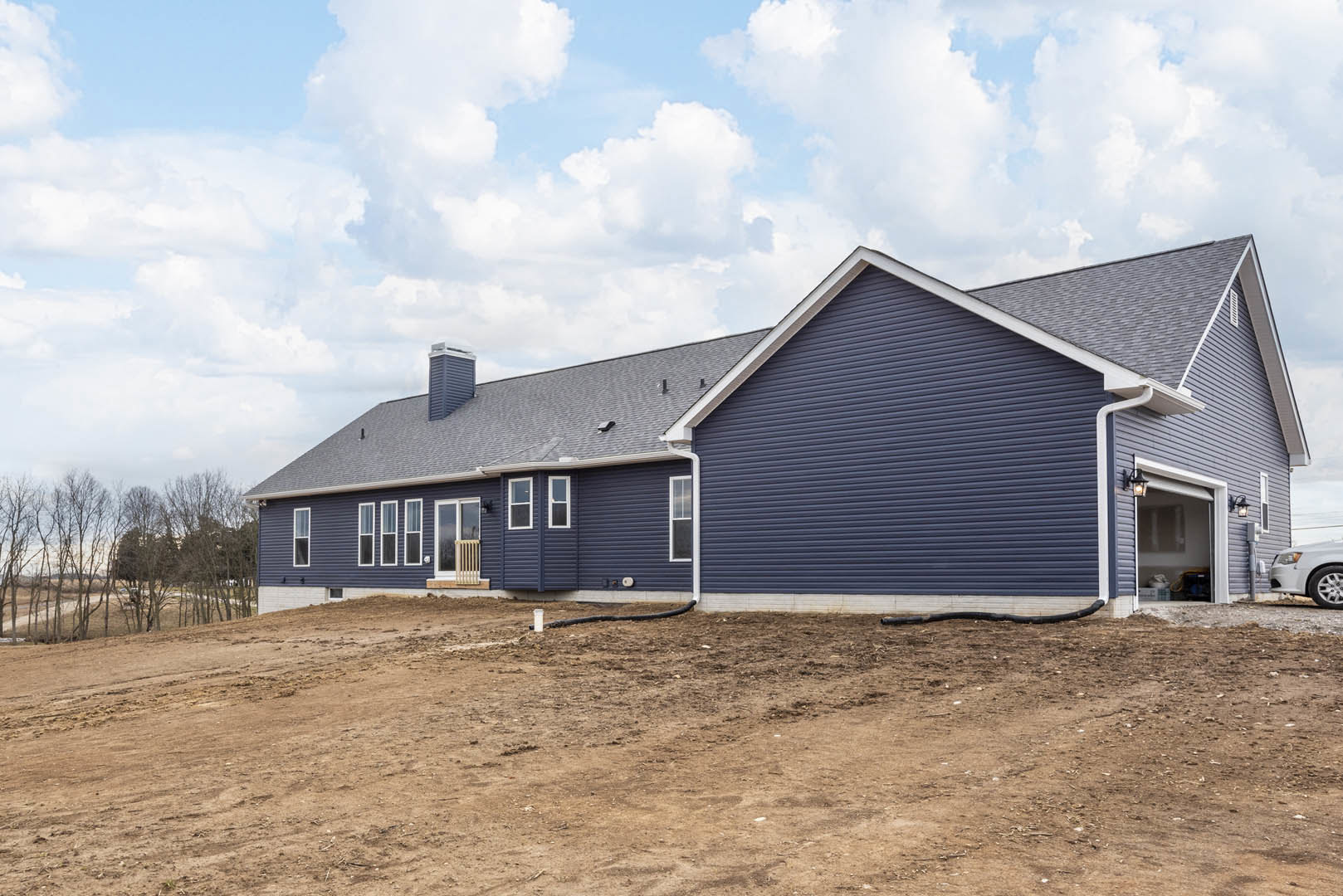 Blue-sided house with blue roof beside dirt hill and exposed pipe, surrounded by trees and open field, illuminated window visible.