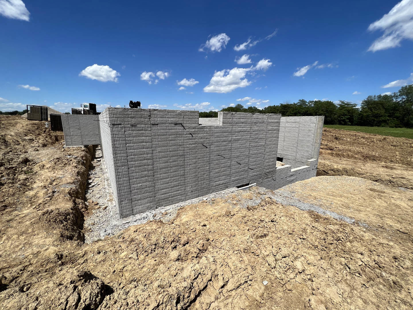 Partially built brick exterior walls on a construction site with exposed soil, scattered construction materials, and a backdrop of trees under a blue sky with white clouds
