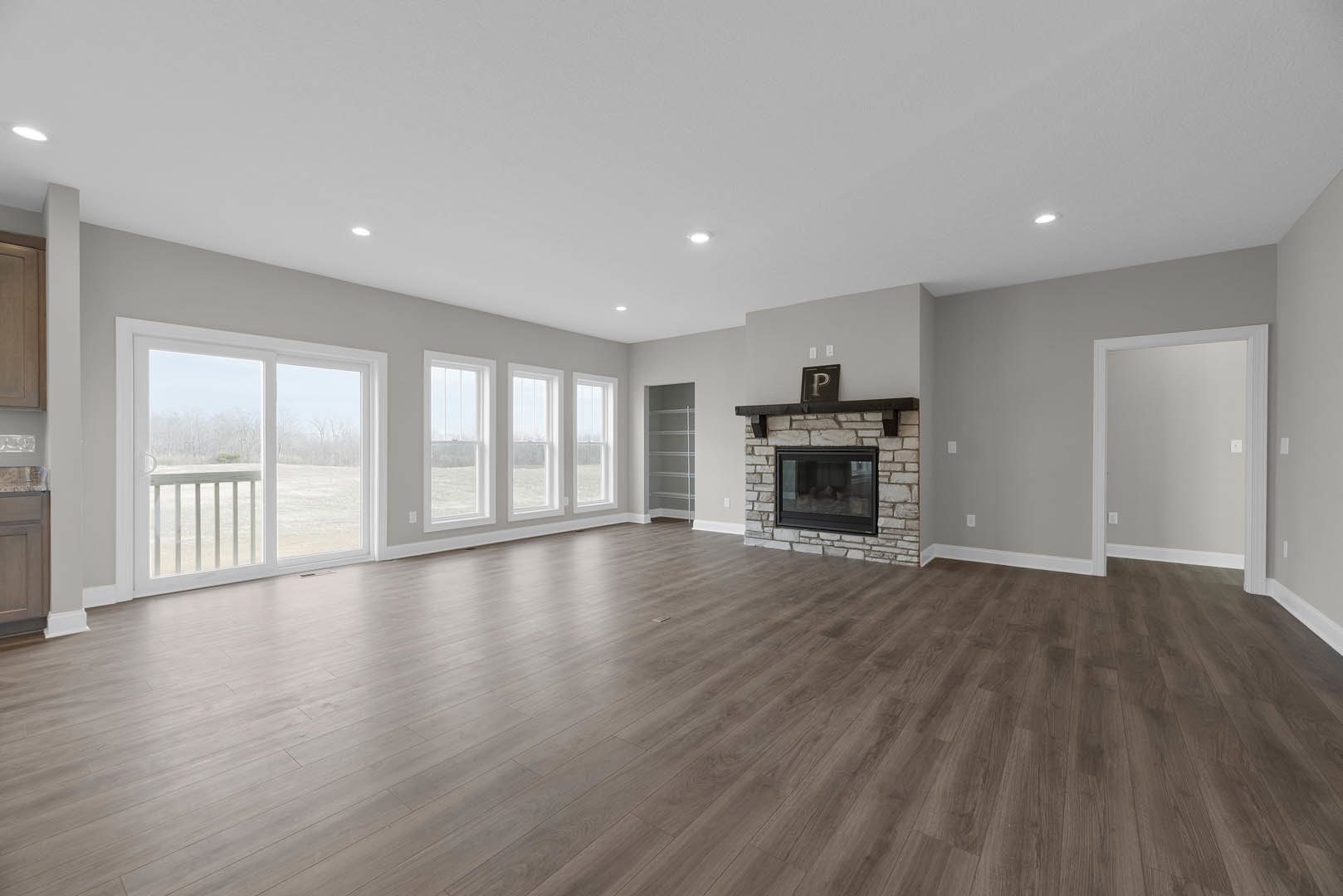 Spacious room with hardwood flooring, brick accent wall featuring a window, white framed door, sliding glass door overlooking a field, and central fireplace.