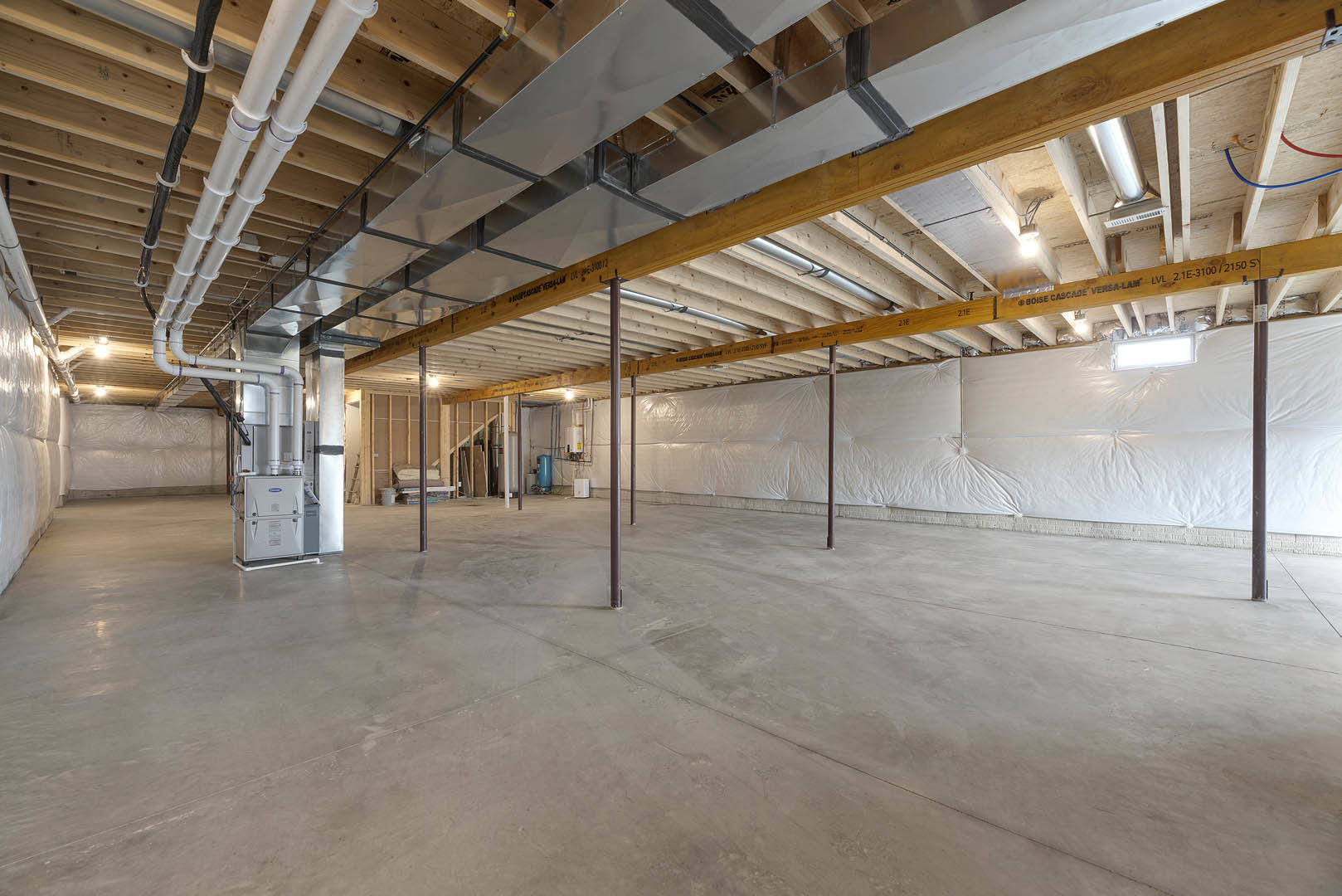 Spacious unfinished room with concrete floor, exposed metal beams and pipes on ceiling, white wall partially covered by a sheet, and a white rectangular box with blue label.
