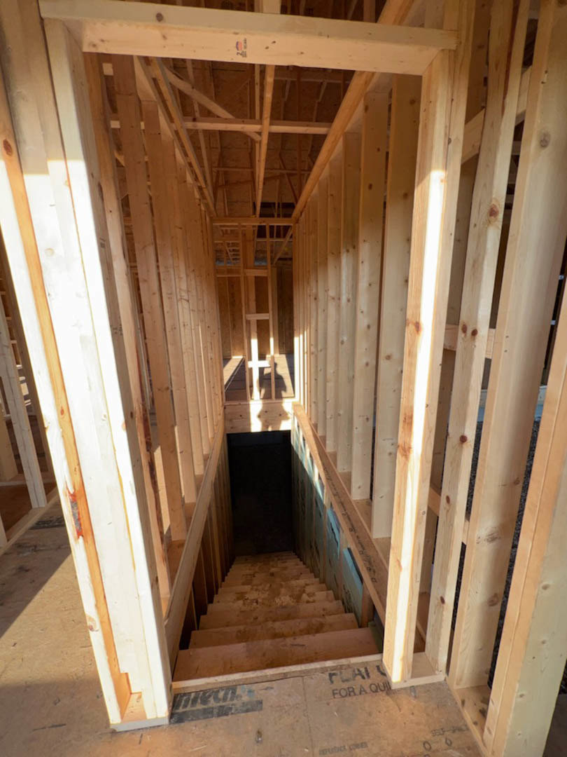 Exposed wooden framing and beams inside a house under construction, unfinished staircase, black wall panel, and construction materials with visible logo