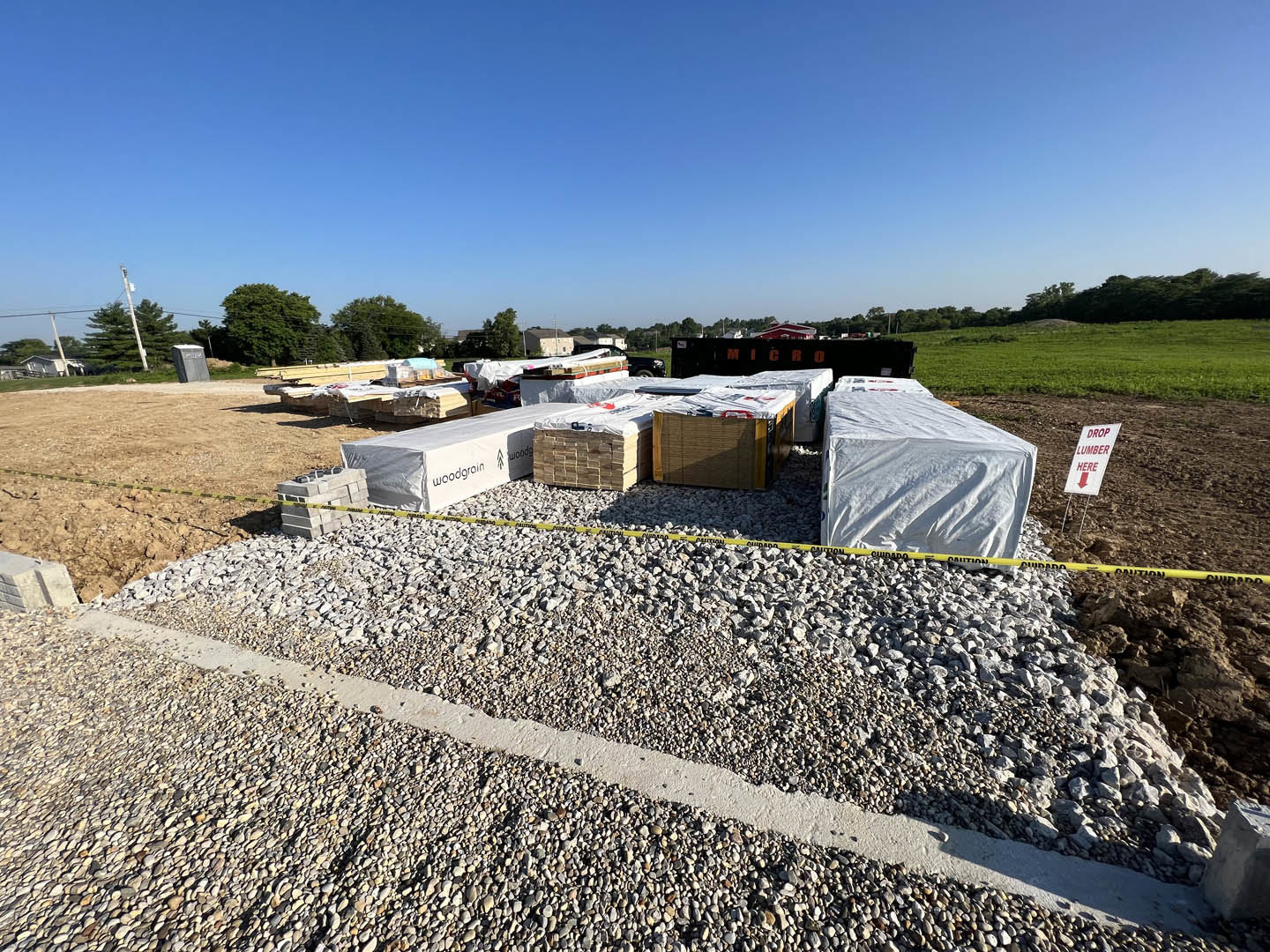 Construction site with stacked pallets, piles of rocks, white plastic coverings, white tarp over a truck, black and white signs with red lettering, grassy field and blue sky in