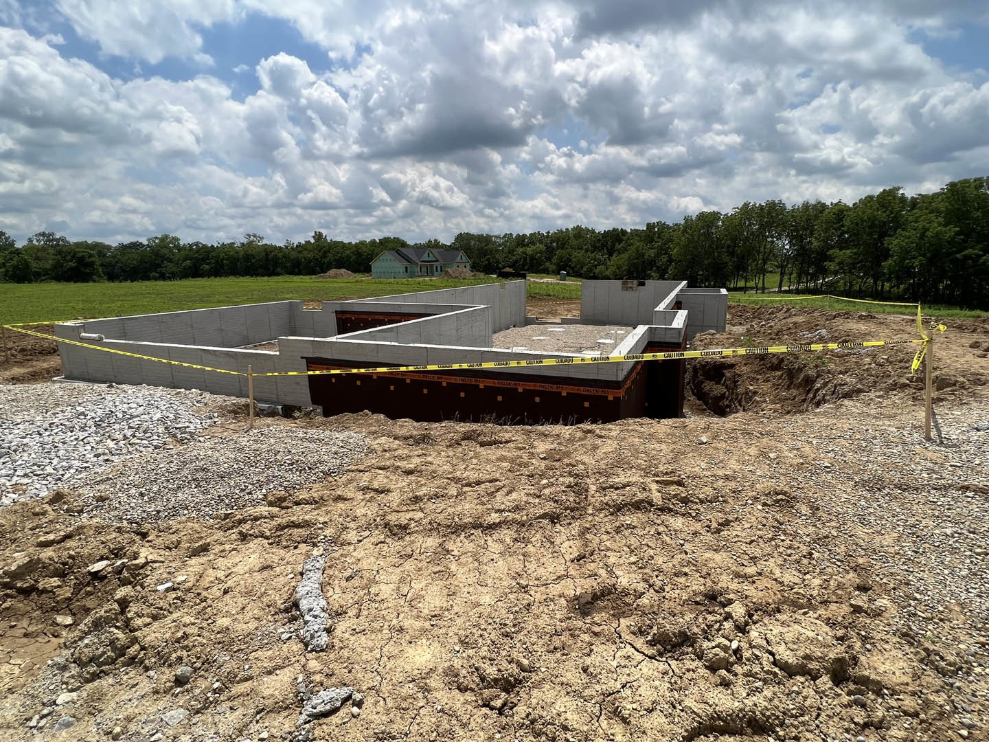 Concrete house foundation surrounded by dirt and rocks, yellow caution tape marking the construction area, cloudy sky overhead, grassy patches and trees in the background