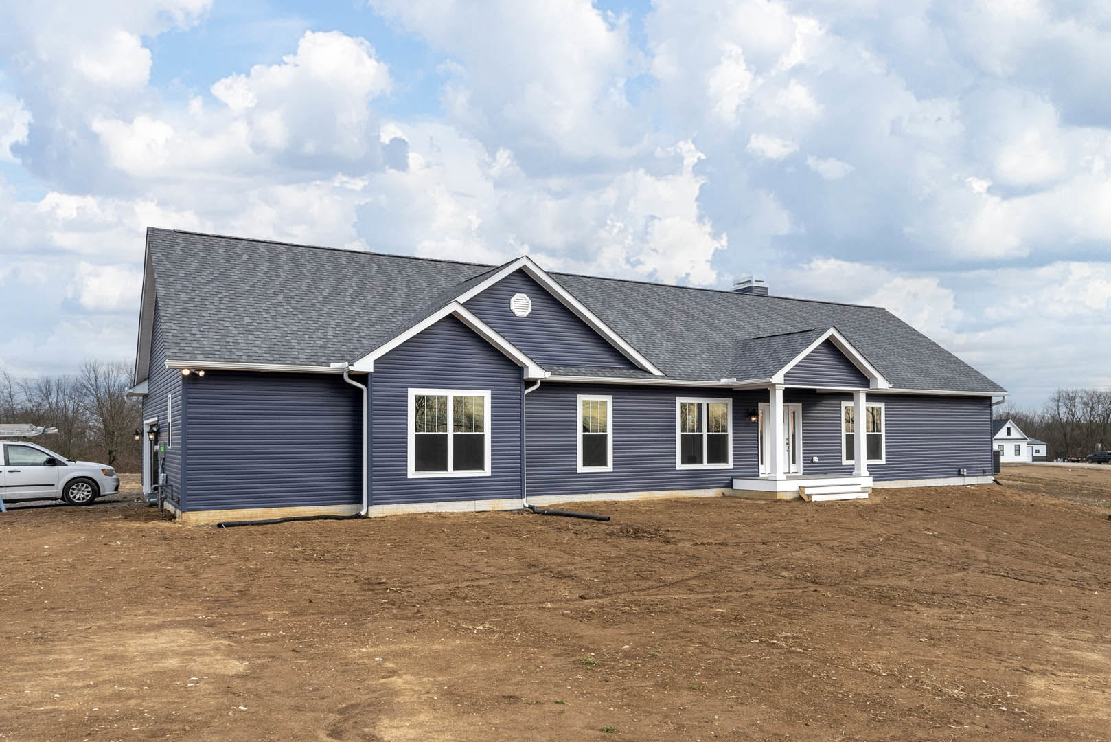 Blue siding house with white-framed windows, dirt field in front, exposed pipe near foundation, white car parked roadside, cloudy sky overhead