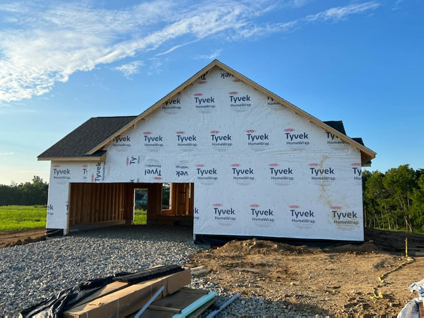 White-walled house under construction with exposed roof framing, dirt and rocks in foreground, trees in background under sunny sky