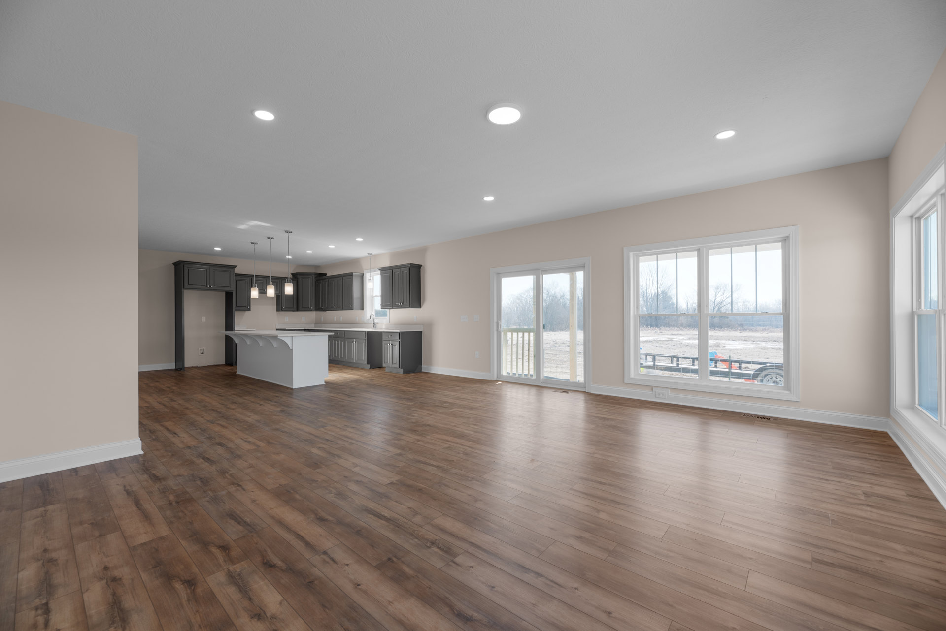 Spacious open-plan room with hardwood flooring, white kitchen island, glass-paneled door, and large window overlooking a field and trees