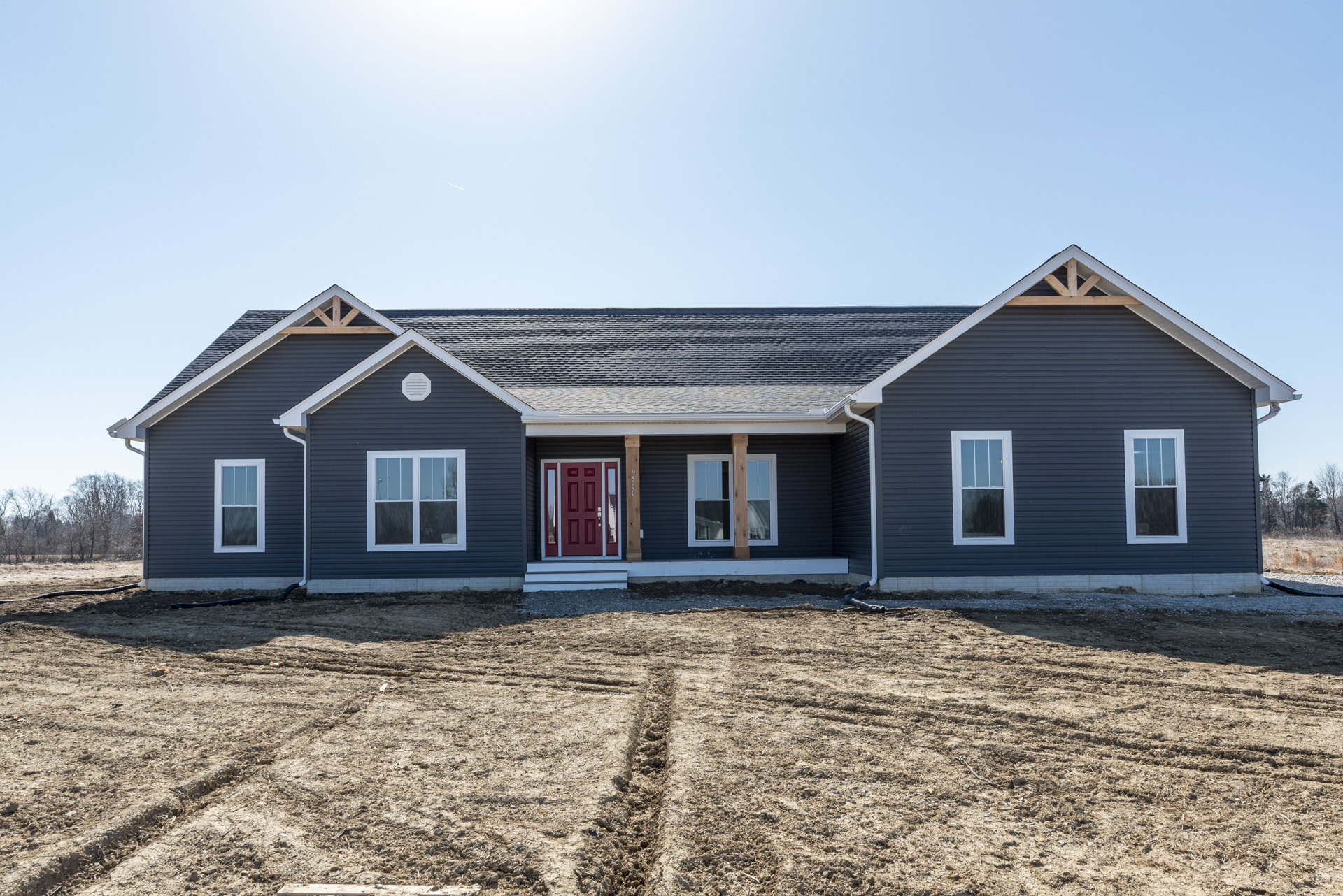 Partially built house with gray siding, red door with glass panes, white-framed window, dirt yard with tire tracks