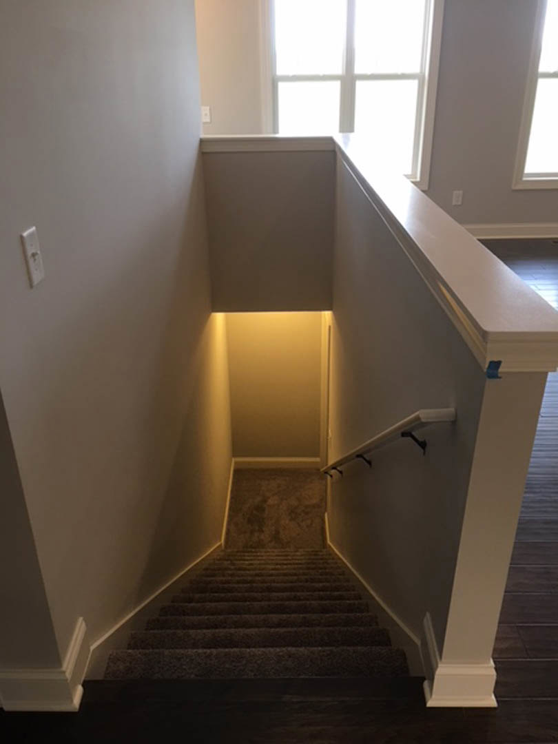 Hardwood staircase with white plaster walls, white railing, and a modern wall-mounted light fixture near the top; natural daylight streams through a nearby window.