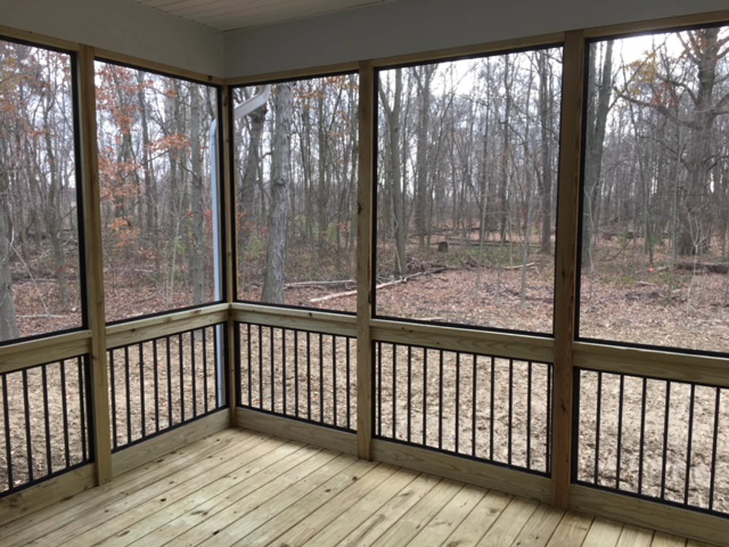 Living room with large black-framed windows overlooking a forest, white walls, wooden floors, and a wooden deck with metal railing visible outside