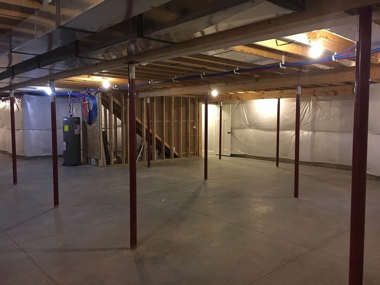 Concrete basement floor with metal support poles, exposed ceiling beams, visible pipes including a close-up blue pipe, and a black electrical box with a yellow sticker.