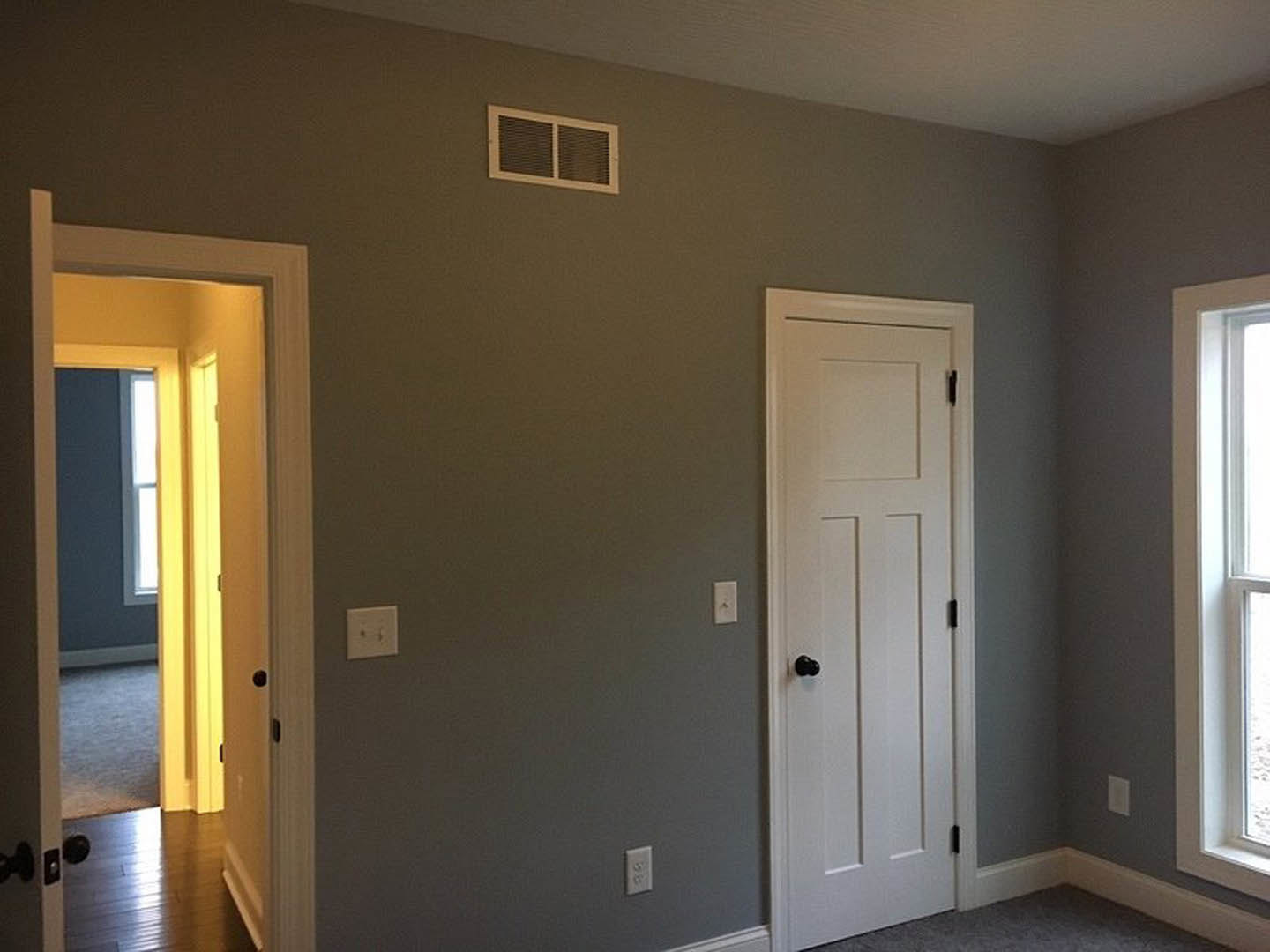 White-painted room with two paneled doors featuring black knobs, wall vent, light switch, and sunlight streaming through doorway onto neutral flooring.