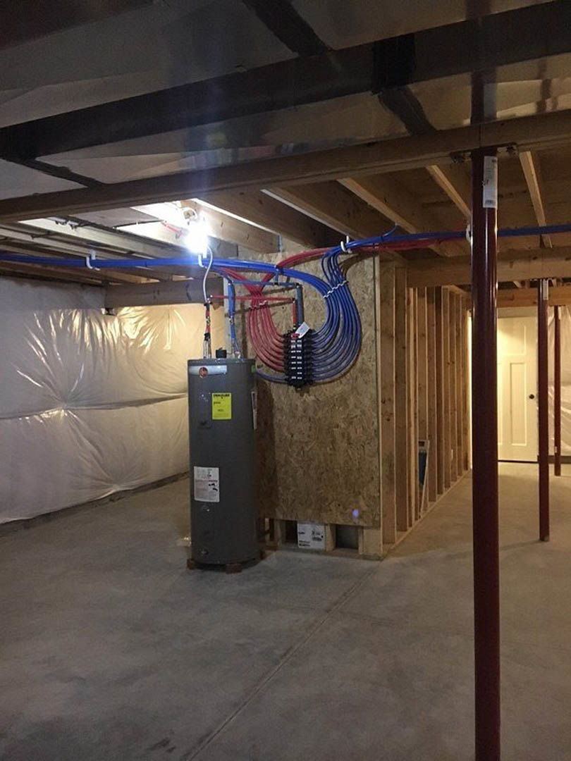 Black cylindrical water heater with connected pipes in a basement, white plastic wall covering, white sign with black text, white door with black knob, exposed ceiling and concrete