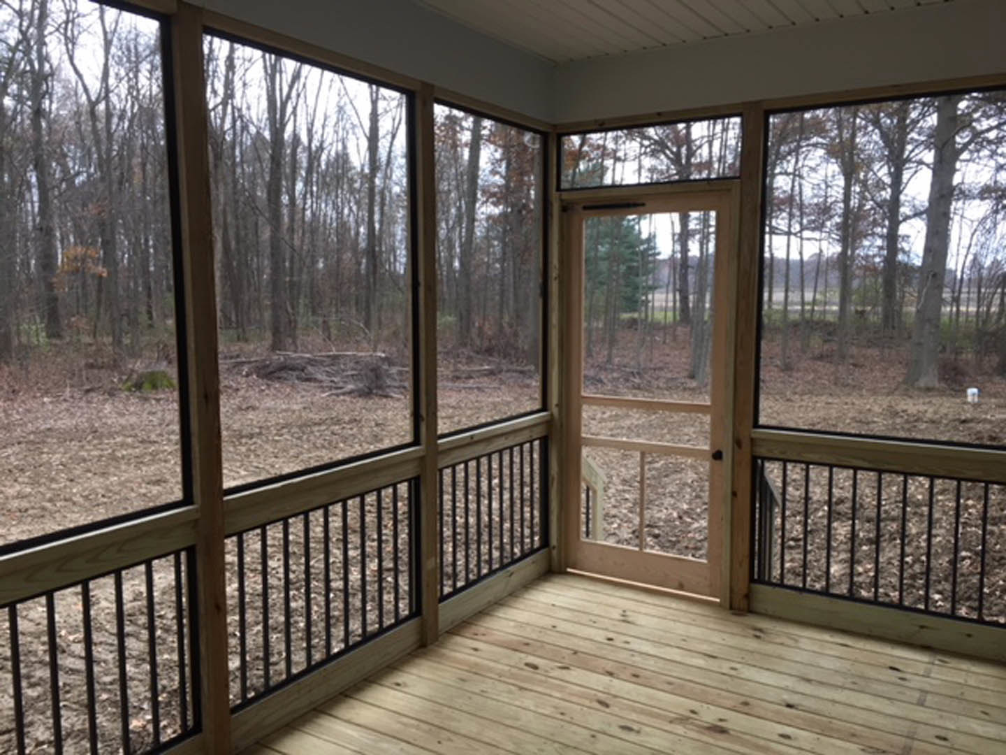 Wood-floored porch with screen door and large windows overlooking dense forest, wooden railing in foreground, trees visible through glass