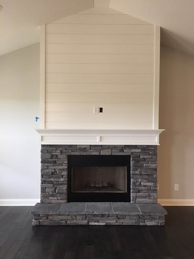 White-paneled wall above a black-framed fireplace with glass window, dark wood floor, stone steps, and brick hearth in a residential living room.