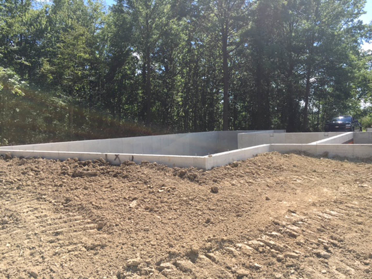 Concrete house foundation set on dirt ground, surrounded by trees in a wooded area, with a cement block wall and a parked car partially visible in the background