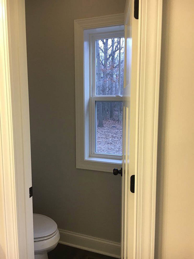 Modern bathroom featuring a white toilet, black floor with white trim, window overlooking trees, and a white door with chrome handle