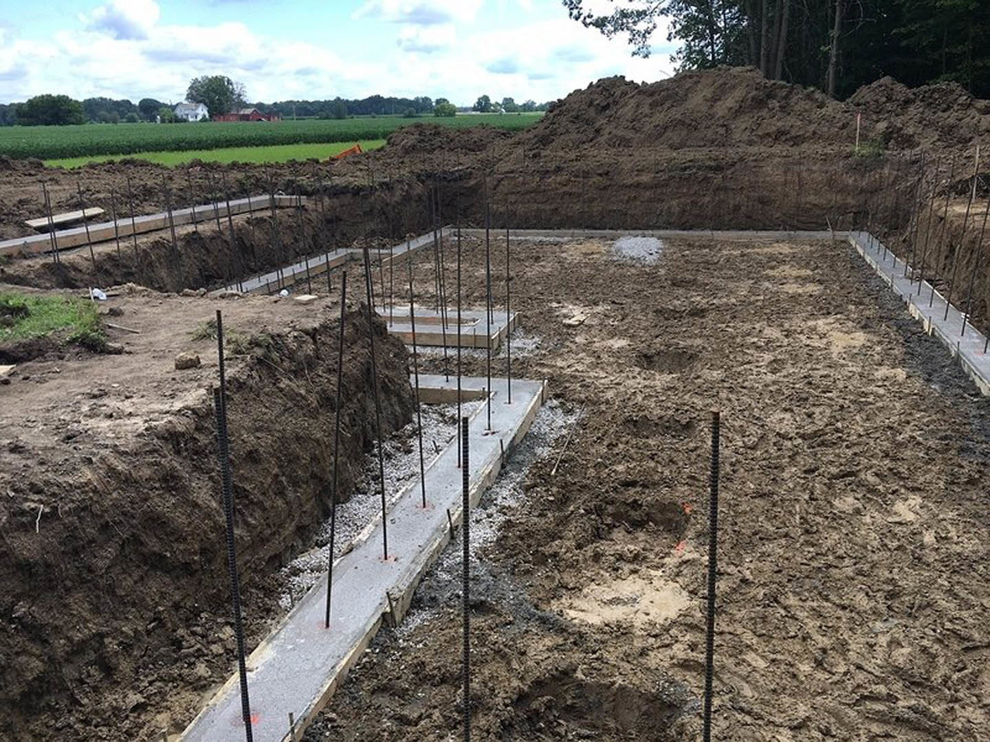 Concrete foundation slab with exposed metal rebar set in brown soil, surrounded by dirt hills and leafy trees under a partly cloudy sky